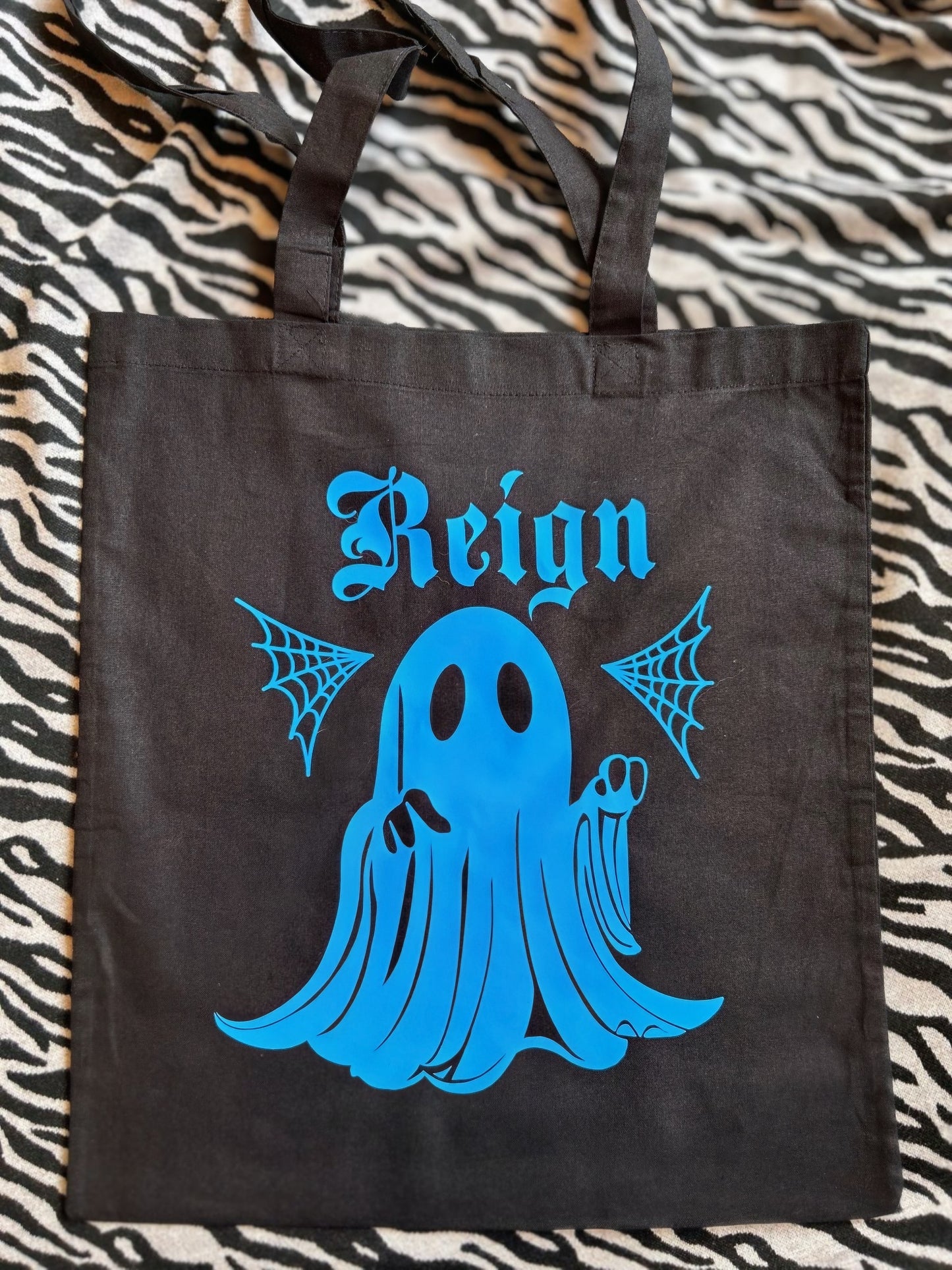 A tote bag, black with a print of a ghost, in blue with a name. This is an example of a customisation. Displayed on zebra print background