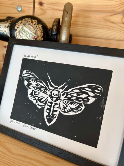 Original lino print of a death moth on black paper, displayed in a frame, with artistic tools in the background.